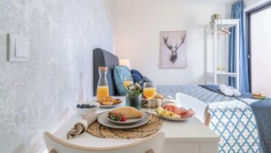 A neatly arranged breakfast table with a variety of foods and drinks, a bed with a patterned bedspread, a wall-mounted shelf with a plant, and a deer head artwork on the wall.
