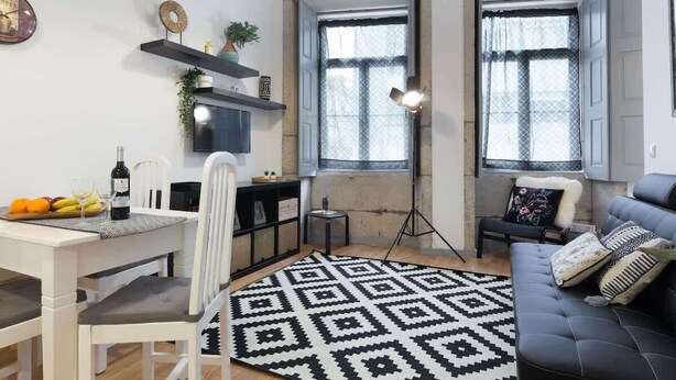 Uma sala de estar moderna com um sofá, uma mesa de jantar com cadeiras, um tapete estampado e duas janelas com cortinas.
