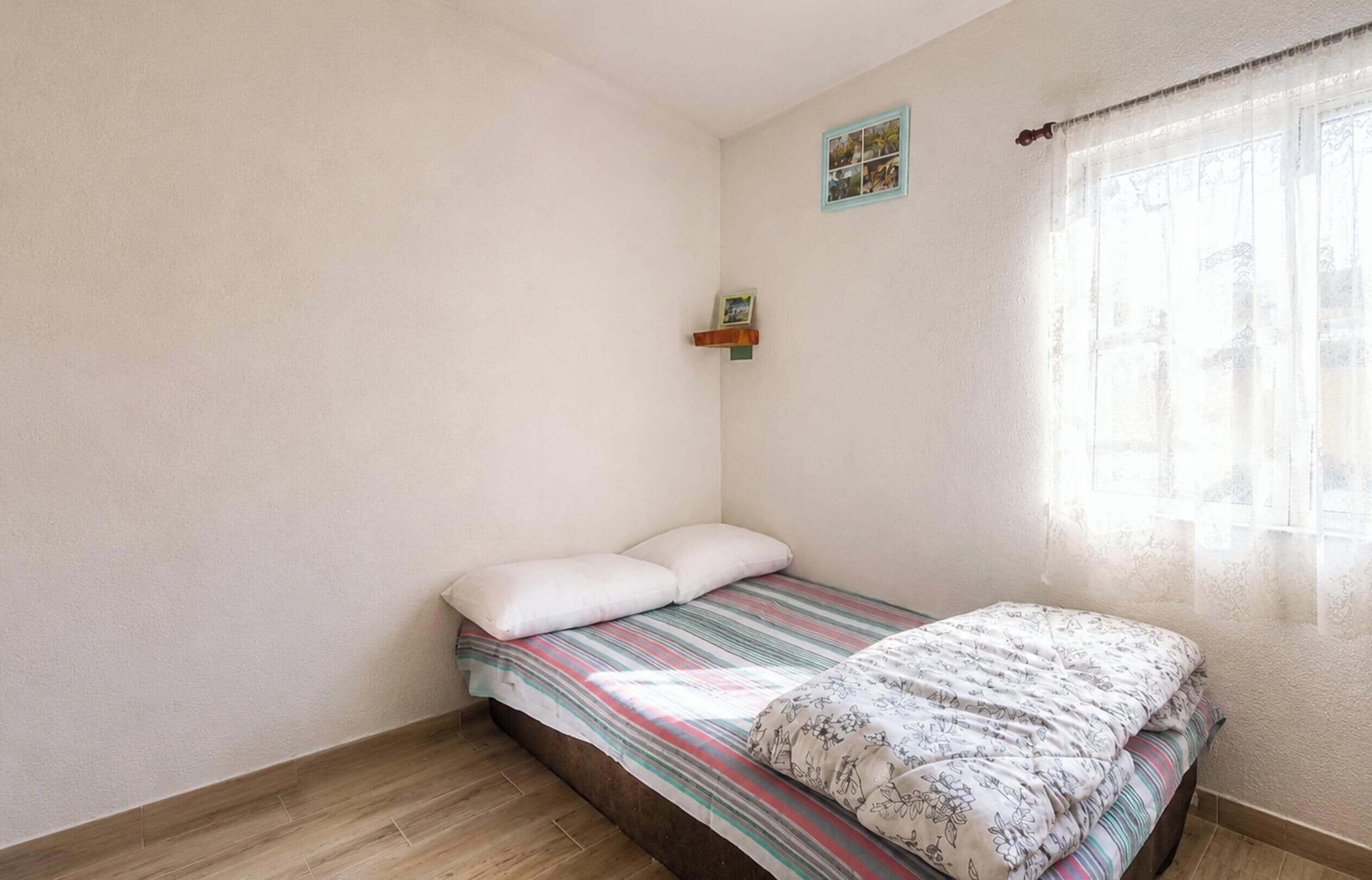 Una habitación pequeña con cama individual, una colcha a rayas, una almohada y una pequeña estantería con un cuadro.