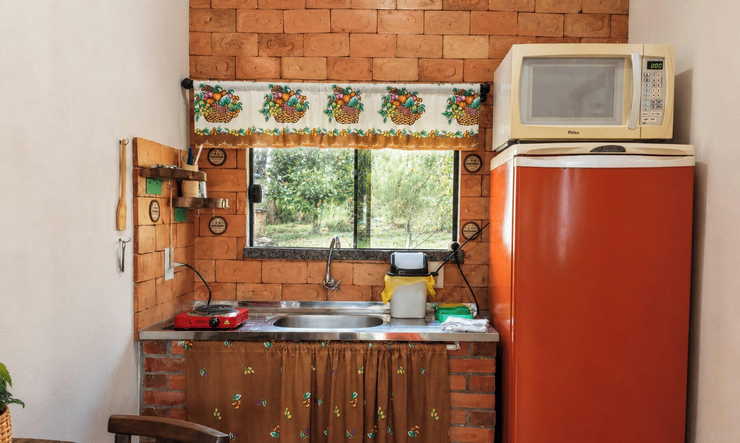 Une cuisine compacte avec un mur en briques, une fenêtre ornée d’un rideau représentant un panier de fruits, un réfrigérateur rouge et un four à micro-ondes posé sur le dessus.