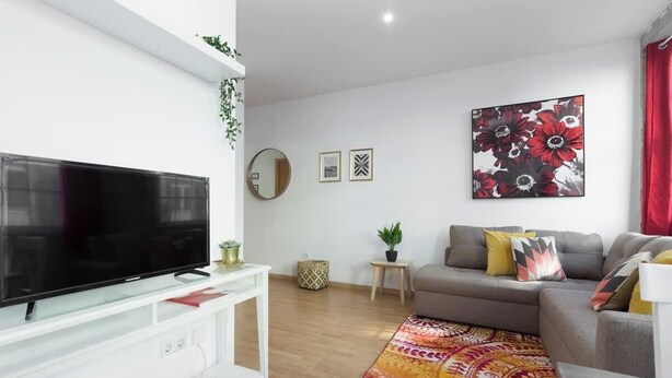 A modern living room with a flat-screen TV, a grey sofa, a colorful rug, and a large floral artwork on the wall.
