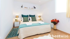 A bedroom with a bed, bedside tables, a nightstand, a wall shelf with decorative items, and a window.