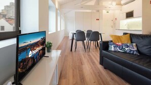 Une chambre d’hôtel moderne avec un plancher en bois, un canapé en cuir noir, un téléviseur sur le côté gauche et un coin repas à l’arrière-plan.
