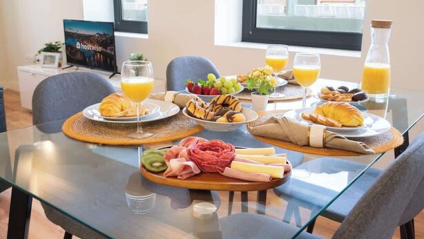 Una mesa de desayuno con diversos alimentos y bebidas, entre ellos frutas, queso, embutidos y cruasanes, con vistas a la ciudad al fondo.