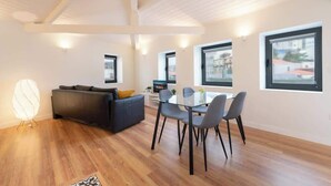Una sala de estar moderna con un sofá oscuro, una mesa de comedor de cristal con sillas y grandes ventanales con vistas a edificios.