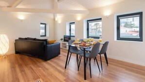 Una sala de estar moderna con un sofá oscuro, una mesa de comedor con sillas y ventanas amplias con vistas a la ciudad.