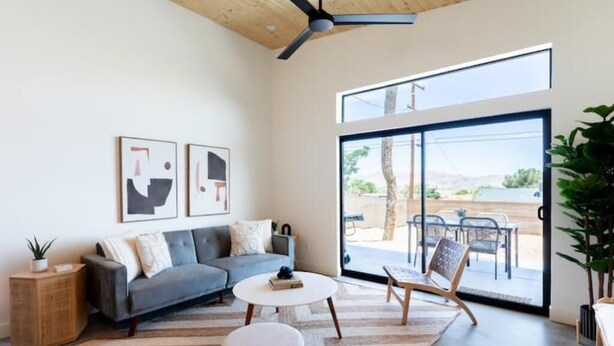 A modern living room with a grey sofa, a round coffee table, and a large sliding glass door leading to an outdoor patio. - Modern Joshua Tree Village Home (Joshua Tree)