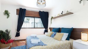 A neatly arranged bedroom with a bed, bedside tables, a nightstand, a window with curtains, and a hanging plant.