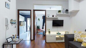 A modern living room with a sofa, a flat-screen TV, and a small table with a decorative bowl. A hallway leads to a kitchen area with cabinets and a refrigerator.