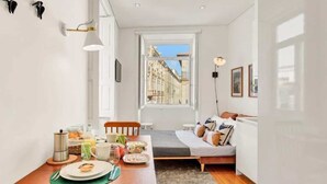 Una habitación compacta con una mesa de comedor dispuesta para el desayuno, una cama con almohadas, un ventanal con vista a la ciudad y una pequeña zona de kitchenette.