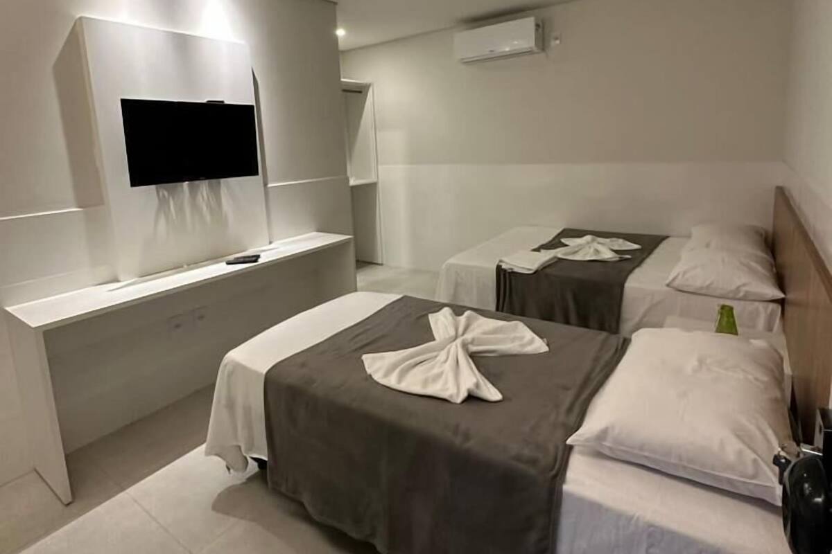 Ein modernes Hotelzimmer mit einem an der Wand befestigten Flachbildfernseher, einem Nachttisch, zwei Betten mit weißer und grauer Bettwäsche und einem kleinen Nachttisch mit einer grünen Flasche.