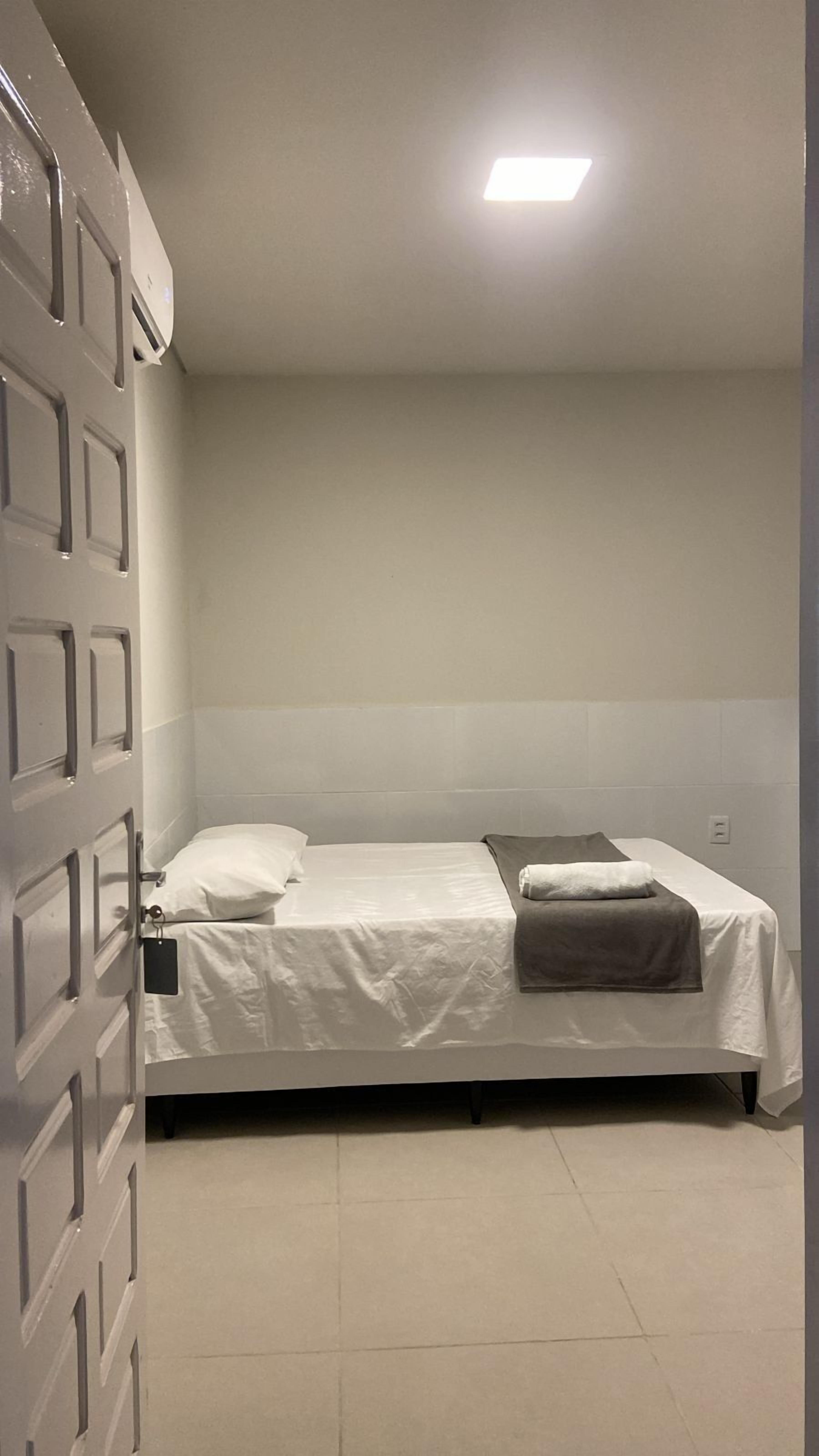 Une petite chambre avec un lit simple, recouverte d’une housse de couette blanche unie, d’une couverture grise et d’un oreiller blanc. La chambre présente un aménagement simple et fonctionnel, avec un mur uni et un éclairage au plafond.
