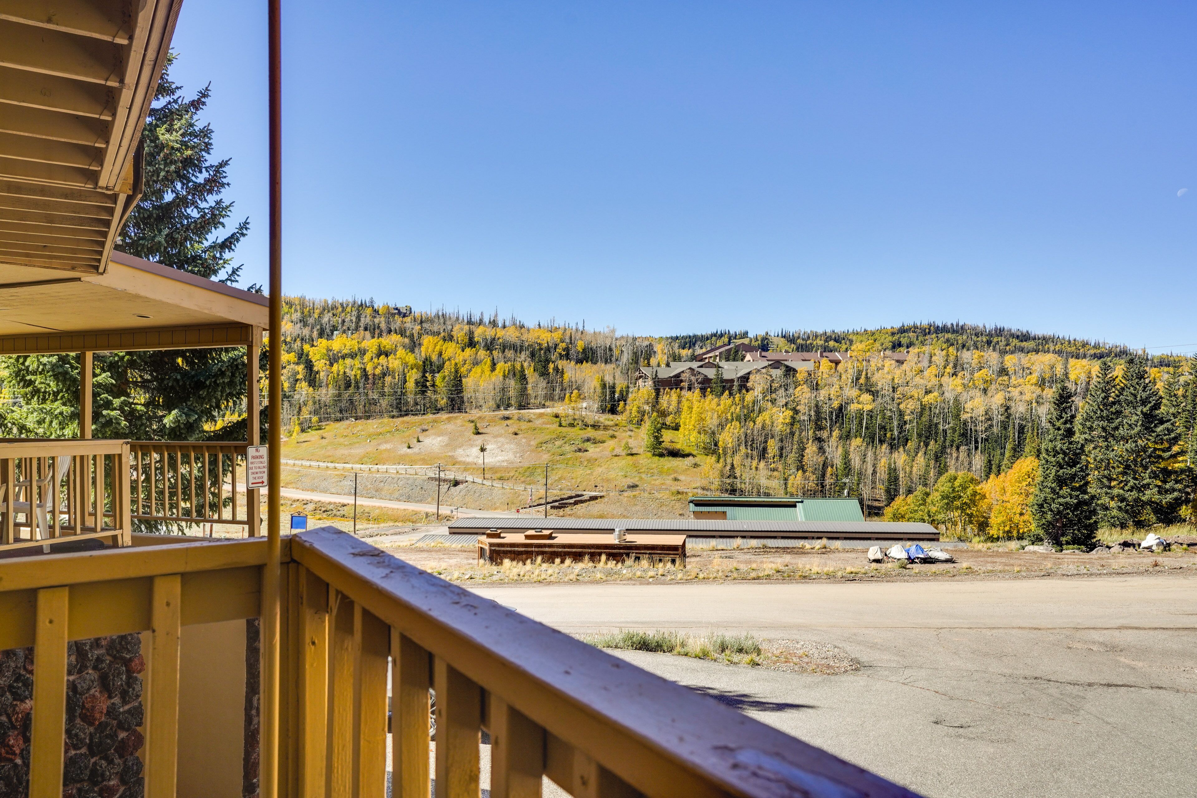 A view from a wooden balcony overlooking a parking area and a forested hillside with autumn-colored trees.