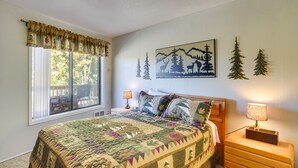 A bedroom with a large bed, a nightstand with a lamp, a window with curtains, and a wall painting of trees. - Cozy Condo w/ Fireplace - Near Brian Head Resort! (Brian Head)