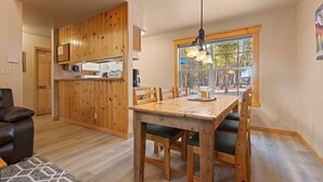 A wooden dining table with chairs, a kitchen area with wooden cabinets, and a large window with a view of trees.