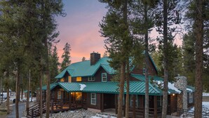 A rustic lodge with a green metal roof, wooden exterior, and stone chimney, surrounded by tall pine trees.