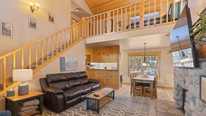 A spacious living area with a leather sofa, a wooden coffee table, and a stone-patterned rug. A wooden staircase leads to an upper level, and a stone fireplace is visible on the right.