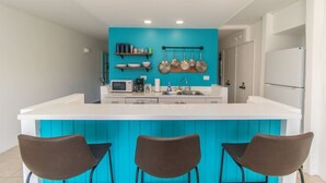 Une cuisine moderne dotée d’un îlot central, de plans de travail blancs et de meubles de couleur turquoise. On y trouve trois chaises marron autour de l’îlot, un four à micro-ondes et une machine à café sur le plan de travail.