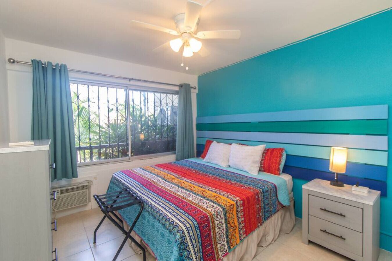 A bedroom with a large bed, a nightstand, a ceiling fan, a window with curtains, and a view of greenery outside.