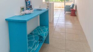 Un couloir avec un banc bleu, une petite table sur laquelle sont posés divers objets, et une vue sur un espace extérieur comprenant une chaise et une petite table.