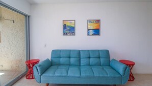 Un canapé de couleur turquoise dans une pièce aux murs blancs, avec deux petites tables d’appoint rouges et deux affiches encadrées accrochées au mur.