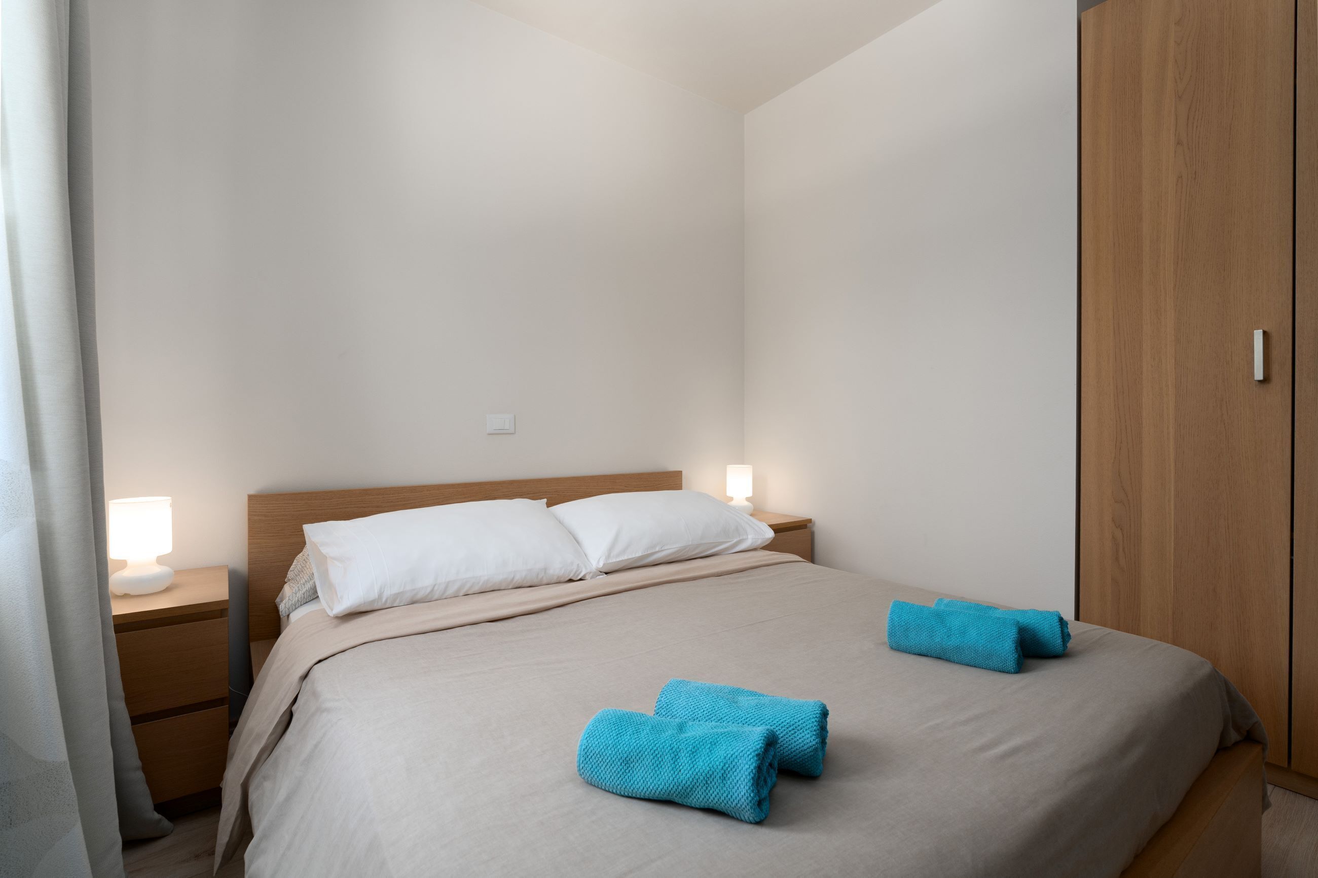 Ein ordentlich bezogenes Bett mit zwei blauen Handtüchern, einem hölzernen Kopfteil, einem Nachttisch mit Lampe und einem Kleiderschrank.