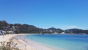A beach with clear blue water, sandy shore, and buildings in the background. - Silver Sands, 29 Victoria Pde water views and across the road to the water (Nelson Bay)