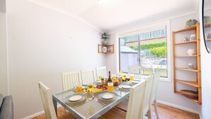A dining area with a glass table, white chairs, and a view of the outdoors through large windows. - Silver Sands, 29B Victoria Pde - across the road to the water (Nelson Bay)