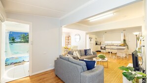 A modern living room with a grey sofa, a coffee table, and a dining area with a wooden table and chairs. A kitchen with white cabinets and a dining table is visible in the background. - Silver Sands, 29B Victoria Pde - across the road to the water (Nelson Bay)