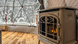 Ein gemütliches Zimmer mit Kaminofen, einer gesteppten Sitzbank und Blick auf schneebedeckte Bäume.