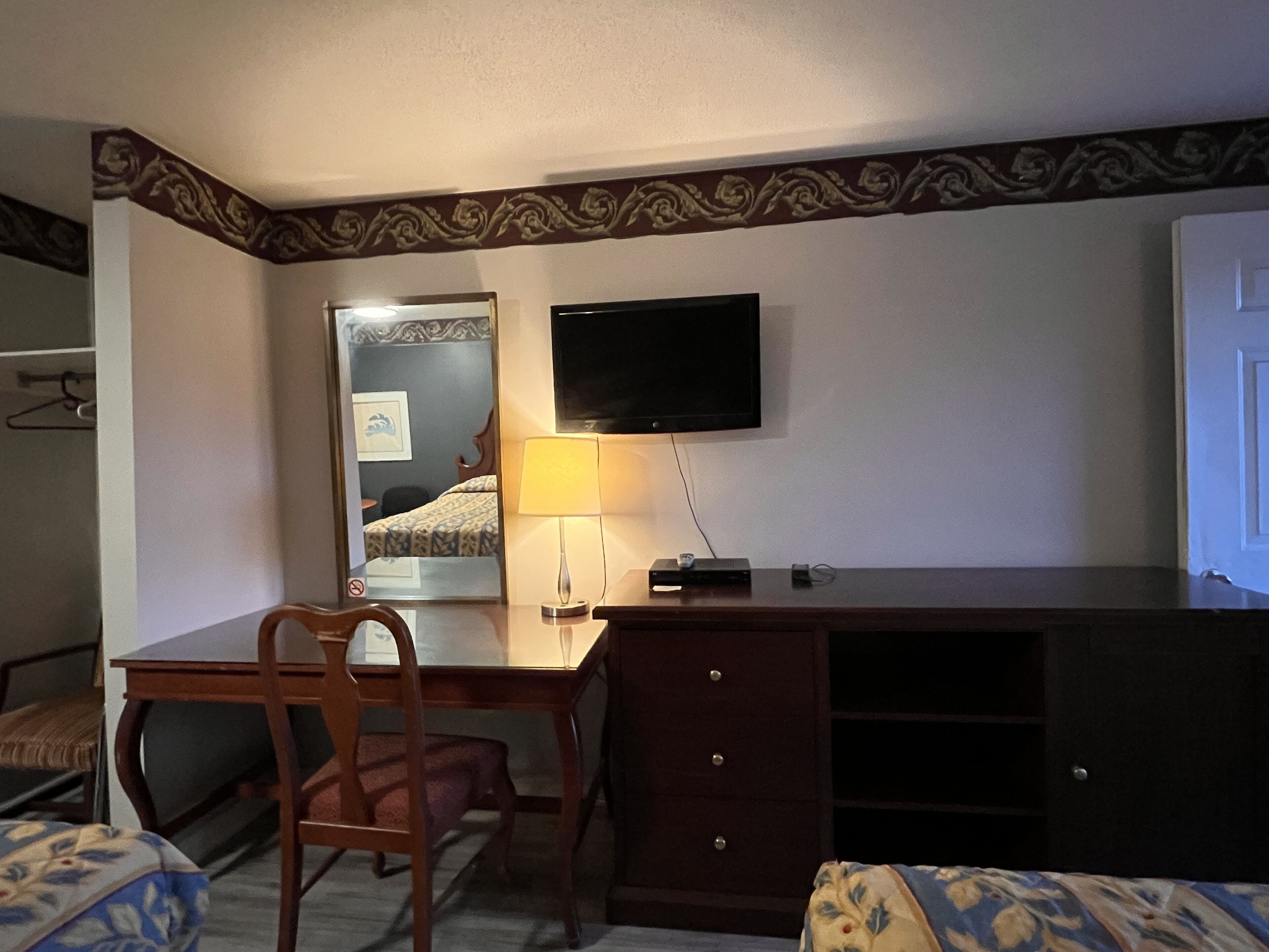 A hotel room with a wooden desk, a chair, a bed with patterned bedding, a flat-screen TV mounted on the wall, and a lamp on the desk.