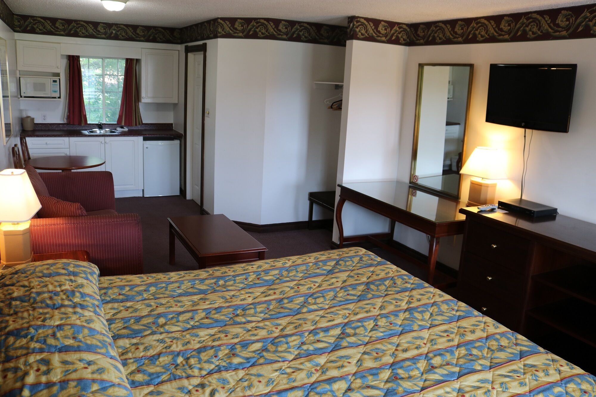 A hotel room with a bed, a desk, a chair, a small table, a mirror, a television, and a kitchenette area.