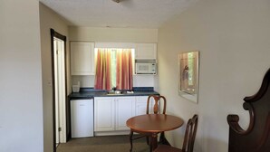 Cocina compacta con armarios blancos, microondas y un refrigerador pequeño. Hay una mesa de comedor con dos sillas y una ventana con cortinas.
