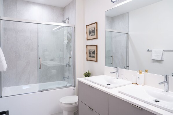 Un bagno moderno con doccia vetrata, due lavandini, un water e quadri appesi alle pareti.