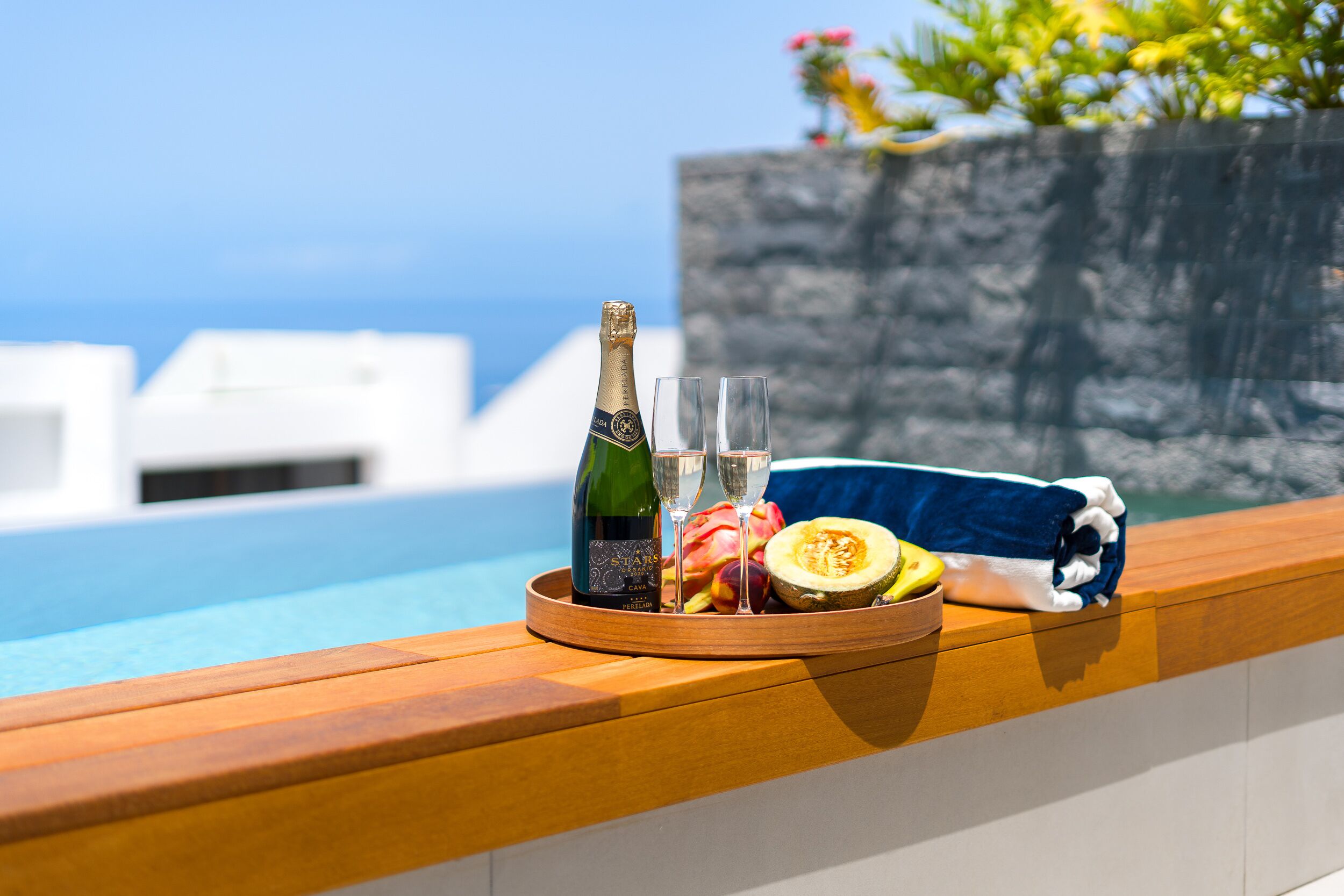 A wooden tray with a bottle of champagne, two glasses, a fruit slice, and a towel by a poolside with a view of the sea.