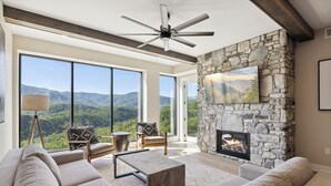 Amplia sala de estar con chimenea de piedra, ventanas grandes con vistas a la montaña y un ventilador de techo.