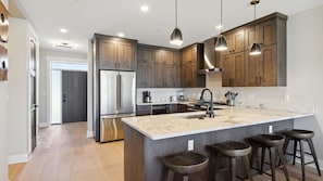 Une cuisine moderne avec des meubles en bois, des appareils électroménagers en acier inoxydable, un grand îlot central avec des tabourets et un éclairage suspendu.