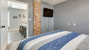Une chambre avec un lit, un mur décoratif en briques, un téléviseur fixé au mur et une porte donnant sur un couloir avec un coin bar.