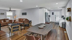 A modern living room with a wooden dining table, leather sofas, and a kitchen area with white cabinets.