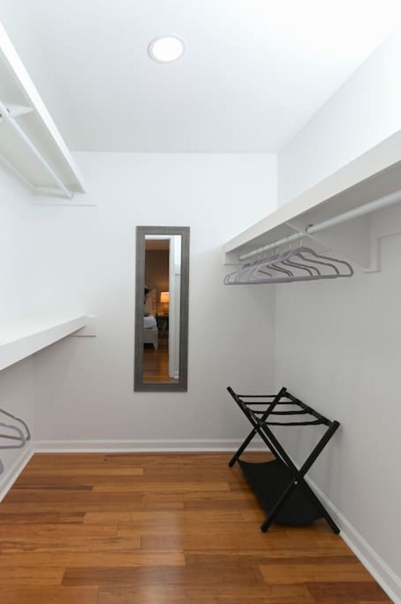 A narrow hallway with wooden flooring, a mirror on the wall, and a black folding chair.