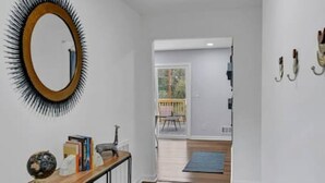 A hallway with wooden flooring, a console table with decorative items, a round mirror with a black frame, and a view into a room with a blue rug.