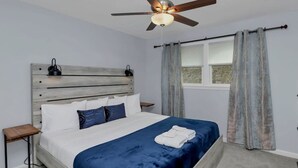 A bedroom with a bed, a ceiling fan, a window with curtains, and a nightstand with a lamp.