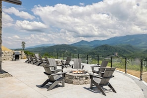 An outdoor seating area with Adirondack chairs around a stone fire pit, overlooking a mountainous landscape.