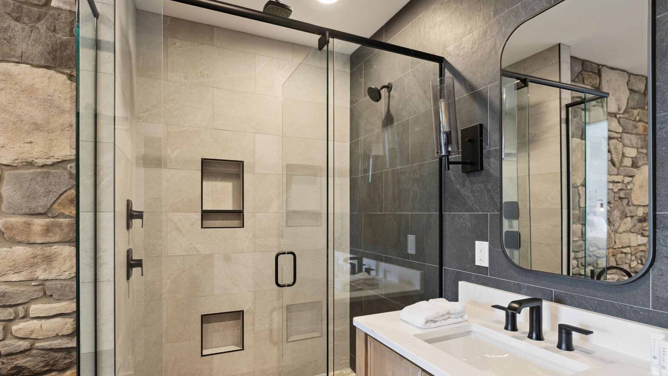 Un baño moderno con ducha de vidrio, un espejo grande y una encimera blanca con dos lavamanos.