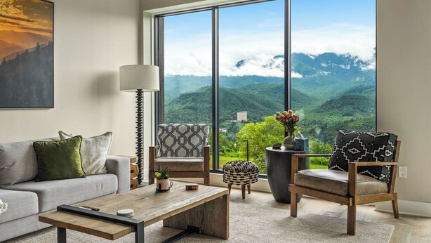 A modern living room with a large window, a sofa, a coffee table, and two patterned chairs. - Stylish Modern 3 BR Condo + Views (Gatlinburg)