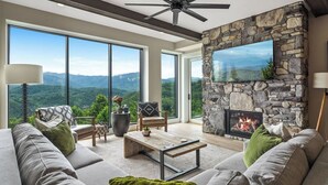 A modern living room with a stone fireplace, large windows, and a ceiling fan. - Stylish Modern 3 BR Condo + Views (Gatlinburg)