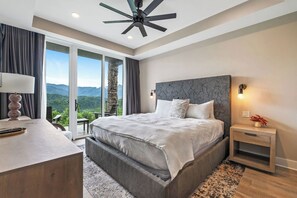 Une chambre avec un grand lit, un bureau, une table de chevet, un ventilateur de plafond et une vue sur les montagnes.