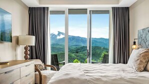 A bedroom with a large bed, a desk, a chair, and a view of mountains through large windows. - Stylish Modern 3 BR Condo + Views (Gatlinburg)