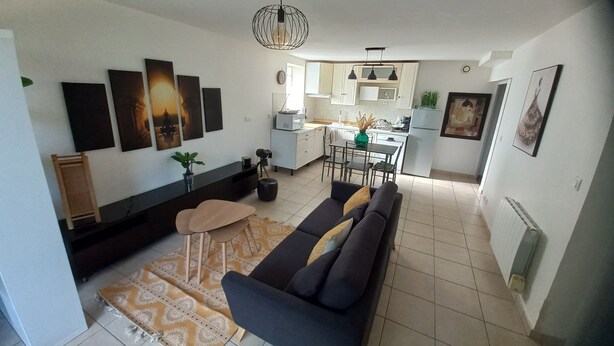A modern living room with a sofa, a coffee table, and a patterned rug. There is a kitchen in the background with cabinets and a dining area. - Appartement T3, Jardin, Parking (Chaponost)