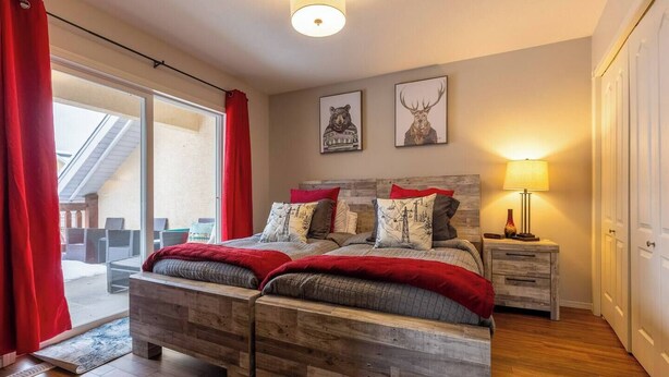 A bedroom with a wooden bed, red curtains, a nightstand, a lamp, and two wall art pieces. - Riverside Chalet + Suite. Enormous Space (Fernie)